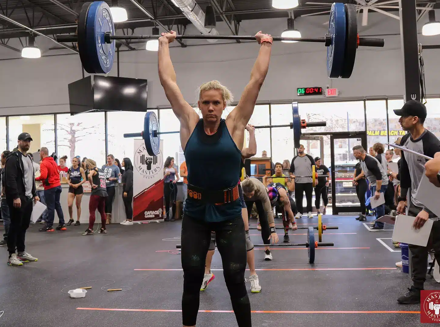 A masters athlete locking out a barbell overhead as the crowd watches